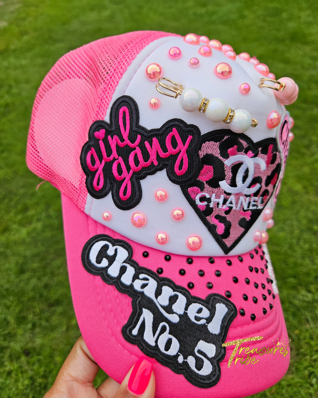 Pink cap with 'girl gang' and 'Cha*el No. 5' text, decorated with pearls on a grass background.